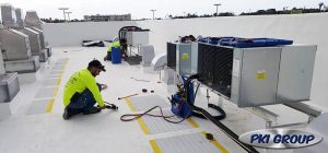 Two technicians in bright yellow shirts working on rooftop HVAC units, surrounded by tools and equipment, with the PKI Group logo in the bottom right corner.