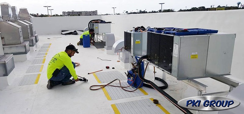 Two technicians in bright yellow shirts working on rooftop HVAC units, surrounded by tools and equipment, with the PKI Group logo in the bottom right corner.