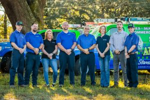 A group of Diamond Air Design team members standing outdoors in front of branded service vehicles, smiling and posed side by side. The team is wearing company uniforms in blue and black, with trees and sunlight in the background.